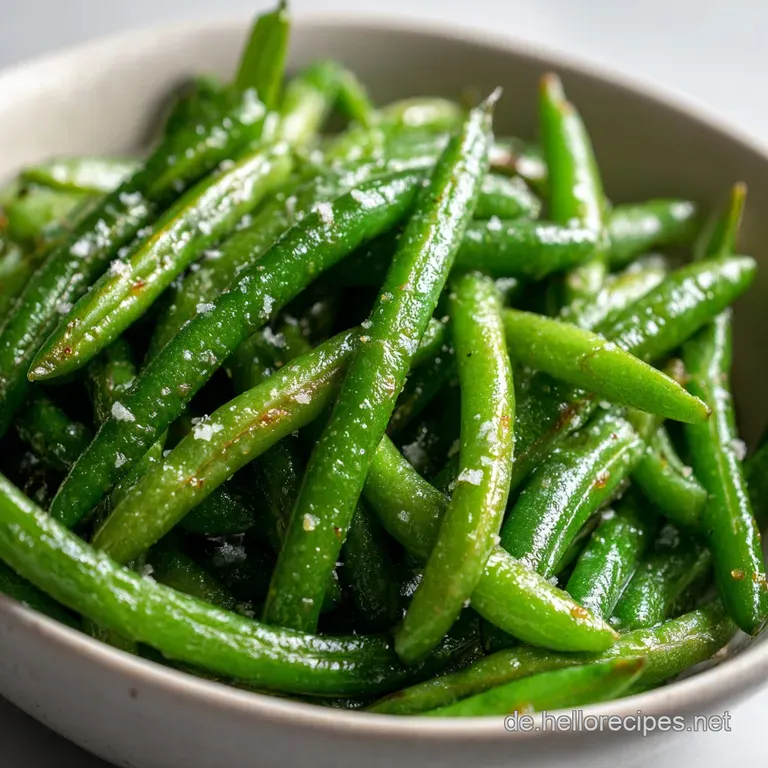 Steaming vibrant green beans served in a shallow white bowl, seasoned, alongside a simple white ceramic spoon.